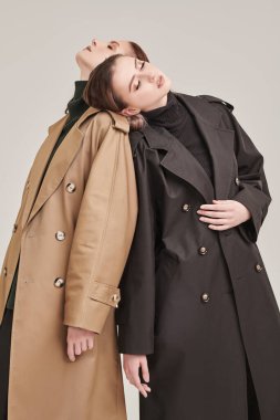 Kadın modası ve güzelliği. İki şık güzel kız model, beyaz bir stüdyonun arka planında moda trençkotlarla poz veriyorlar..