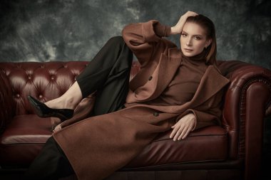 Çekici, kendine güvenen bir kadın deri koltukta poz veriyor moda keçeli ceketi, siyah pantolonu ve yüksek topuklu ayakkabıları var. Kadın modası.
