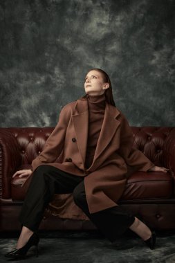 Çekici, kendine güvenen bir kadın deri koltukta poz veriyor moda keçeli ceketi, siyah pantolonu ve yüksek topuklu ayakkabıları var. Kadın modası.