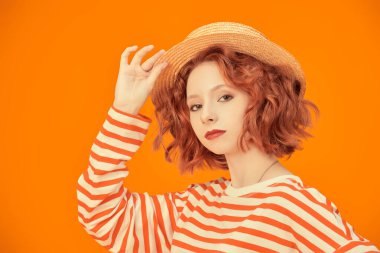 Kıvırcık kızıl saçlı güzel bir kız, hasır şapka giymiş ve sarı stüdyo arka planında turuncu çizgileri olan uzun kollu bir kız. İnsanların duyguları. Yaz modası ve güzelliği.