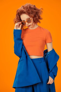 Parlak renkler. Parlak mavi, turuncu giysiler ve güneş gözlükleriyle gülümseyen seksi kıvırcık saçlı kendine güvenen güzel bir kızın portresi. Stüdyo turuncu arka plan.