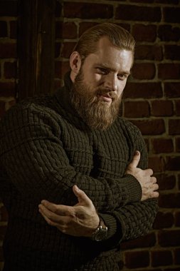 Cesaret ve güç. Tuğla bir duvarın arkasında duran ve sakince ve kendinden emin bir şekilde ileriye bakan örgü örülü süveter giyen iri sakallı bir adamın portresi..