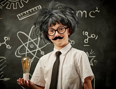 Genç bilim adamları. Gözlüklü, bıyıklı ve peruklu komik çocuk. Küçük profesörün ampulü tuttuğu gibi. Siyah karatahtanın arka planında çizimler var. Eğitim ve erken kalkınma.