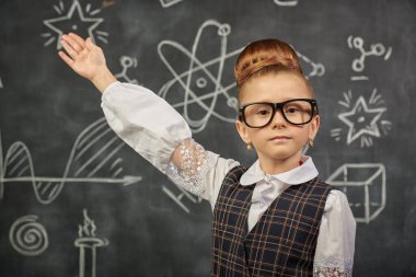 Okul üniformalı ve kameraya ciddi ciddi bakan zeki bir kızın portresi. Bilimsel çizimleri olan siyah bir kara tahta. Genç bilim adamları. Eğitici kavram. 