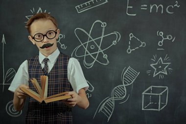 Gözlüklü, bıyıklı, profesör gibi poz veren sevimli bir çocuk. Siyah bir karatahtanın arka planı. Eğitim ve erken gelişim. Genç Bilim Adamları.