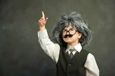 Gözlüklü, bıyıklı ve peruklu şirin bir çocuk. Sanki küçük profesörün bir fikri varmış gibi. Kopya alanı olan siyah bir karatahtanın arka planı. Eğitim. Küçük Bilim Adamı.