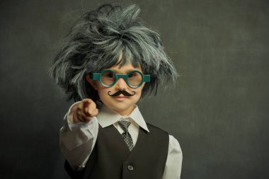 Gözlüklü, bıyıklı ve peruklu şirin bir çocuk. Küçük profesörün kameraya baktığı gibi. Kopya alanı olan siyah bir karatahtanın arka planı. Eğitim. Küçük Bilim Adamı.