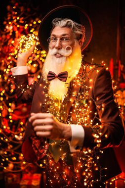 Zarif sakallı ve bıyıklı yakışıklı, modern bir Noel Baba resmi. Şapka takıyor, şık bir takım elbise giyiyor ve altın ışıklarla çevrili gözlükler takıyor. Sihirli Noel ve yeni yıl. Moda.. 
