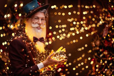 Sihirli Noel ve yeni yıl. Zarif sakallı ve bıyıklı yakışıklı, modern bir Noel Baba resmi. Şapka takıyor, şık bir takım elbise giyiyor ve altın ışıklarla çevrili gözlükler takıyor. Moda.. 