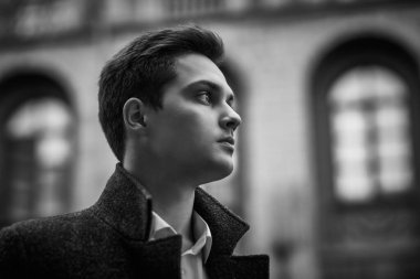 Paltolu yakışıklı, modern bir adam bulutlu bir günde şehir caddesinde duruyor ve ileriye bakıyor. Siyah-beyaz fotoğraf. İnsanlar ve duygular, yaşam tarzı. 