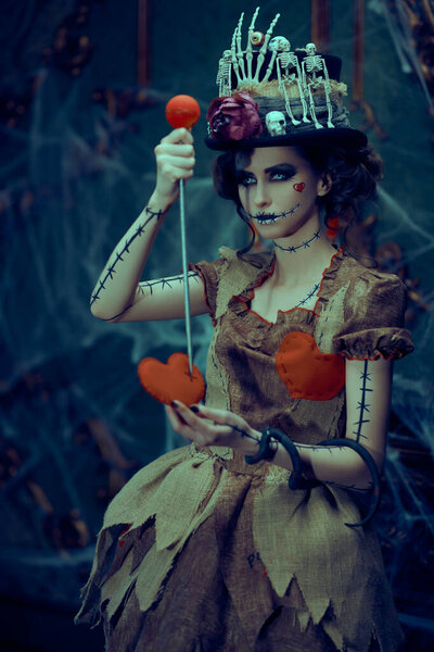 Красивая и вероломная молодая женщина, одетая как кукла вуду, стоит в старом заброшенном замке и втыкает булавку в игрушечное сердце жертвы. Религия вуду. Хэллоуин.