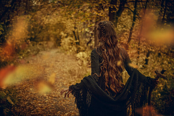 Женщина-шаман в черной одежде с этническими орнаментами на лице стоит в осеннем лесу с посохом шамана в руке. Этническая культура. Шаманские ритуалы.