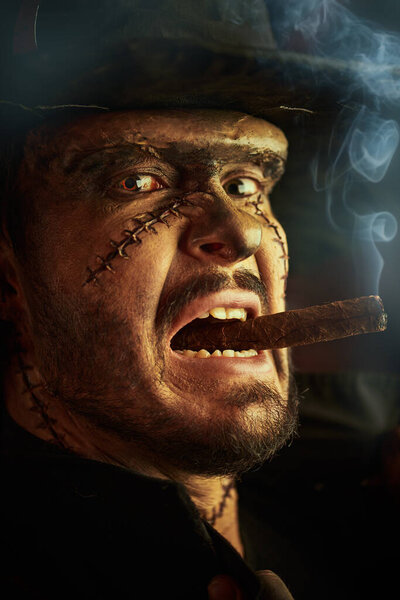 Close-up portrait of a scary man with scars on his face in a black hat, who smokes a cigar, gets angry, and looks with evil eyes. The hero of a horror novel. 19th century style. Halloween.