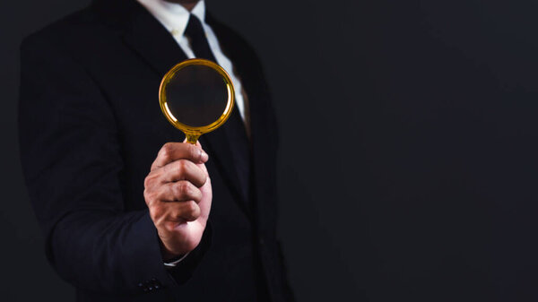 Businessman using magnifying glass against dark background with empty space.