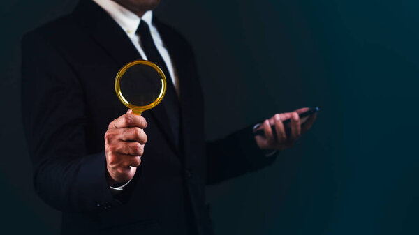 Businessman is using smartphone and magnifying glass searching for something against dark background.