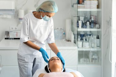 Kadın kozmetik uzmanı müşterinin yüzüne bakıyor ve cildini inceliyor. Mutlu, rahat, genç bir kadın spa salonunda profesyonel yüz bakımı görüyor. 