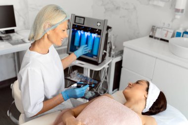 Temizleme prosedürü. Güzel, güzel bir kadın yüzünde temizleme ameliyatı olurken gözleri kapalı yatıyor. Kıdemli doktor gülümseyerek çalışırken ekranı işaret ediyor. 