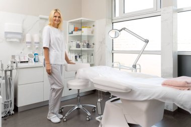 Beyaz laboratuvar önlüklü çekici bir güzellik uzmanı çarşafla kaplı bir şekilde yatağın yanında duruyor. Kameraya bakıyor ve gülümsüyor. 