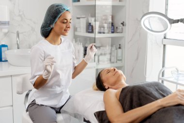 Koruyucu ekipmanlı kozmetik uzmanı kadın, yanında otururken gülümseyerek yaşlı kadın hastasına bakıyor. Kozmetoloji kavramı 