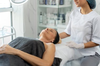 Koruyucu ekipmanlı kozmetik uzmanı kadın, yanında otururken gülümseyerek yaşlı kadın hastasına bakıyor. Kozmetoloji kavramı 