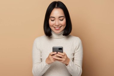 Elinde akıllı telefonuyla gülümseyen iyimser bir kadın erkek arkadaşından gelen romantik mesajlara cevap veriyor. Bej arka planda izole edilmiş kapalı stüdyo çekimleri 