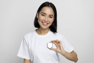 Positive asian woman demonstrating Implantable cardioverter defibrillator (ICDs) at hands while looking at the camera with pleasant smile. Health care and heart concept  clipart