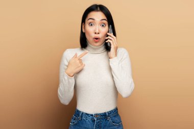 Şaşırmış esmer kız şaşırmış bir ifadeyle cep telefonu kullanıyor iletişim sırasında şok edici haberler duyuyor. Stüdyo çekimi bej arkaplanda izole edildi 