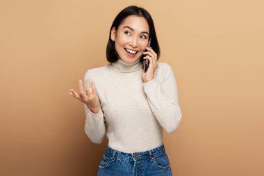Neşeli kadın portresi cep telefonuyla konuşuyor, başka tarafa bakıyor ve neşeyle gülümsüyor, hoş sohbetler ediyor. Bej arka planda izole edilmiş kapalı stüdyo çekimleri 