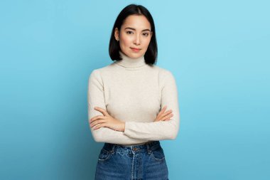 Pozitif yüzlü, kollarını kavuşturarak kameraya bakan güzel bir kadın. Mavi arka planda kapalı stüdyo çekimi 