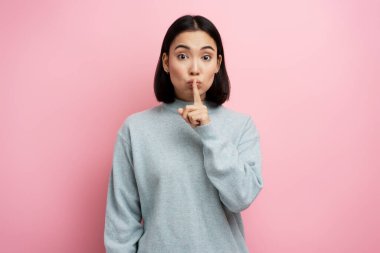 Shh, sessiz ol. Ciddi bir kadın portresi. Ağzında parmaklarıyla sessiz bir jest yapıyor. Sessiz kalmak ve sır tutmak istiyor. Pembe arka planda kapalı stüdyo çekimleri 