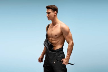 Mavi arka plan üzerinde poz veren güçlü beyaz adam. Çıplak vücutlu kendine güvenen genç bir tamircinin portresi. 