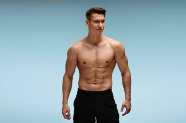 Stüdyoda kameranın önünde mavi arka planda yalnız poz veren yakışıklı, üstsüz, atletik genç adam.