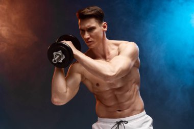 Front image of fit, strong young man with bare torso, training with dumb-bell, showing six pack abs, black background 