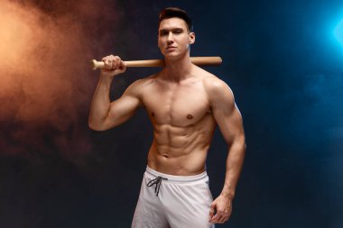 Stüdyoda beyzbol sopasıyla poz veren kaslı bir adamın görüntüsü, kameraya dikkatlice bakıyor. Spor konsepti 