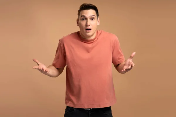Astonished man wearing casual style T-shirt, standing with mouth open in surprise, has shocked expression, hears unbelievable news. Indoor studio shot isolated on beige background