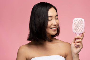 Happy brunette asian woman with silky hair holding modern hair brush isolated over pink colour background. Hair care concept