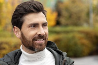 Mature bearded man smiling outdoors while looking away.