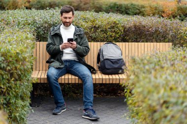 Happy, friendly man is sitting on a bench outdoors. A guy looks at a mobile phone, a backpack is next to him.