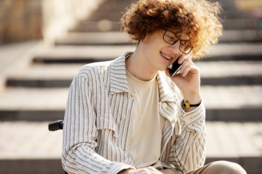 Smiling, young man in glasses in a wheelchair, talking on a mobile phone.