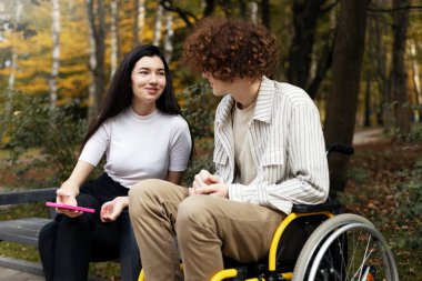 Cheerful, smiling girl is talking to a young guy, he sitting in a wheelchair. The girl holds a smartphone in her hand and sits on a bench on the street.