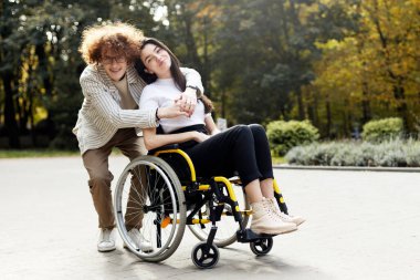 Handsome, smiling, curly-haired man in glasses hugs his girlfriend. A beautiful girl sits in a wheelchair on the street.