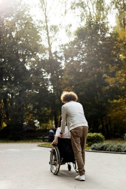 Curly-haired man carries a girl in a wheelchair. Back view, young people are walking on the street.