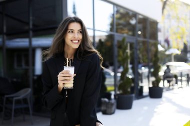 Elinde kahveyle gülümseyen, çekici bir kız sokakta dikilip başka tarafa bakıyor. Kız modern, cam bir binanın arka planında duruyor..