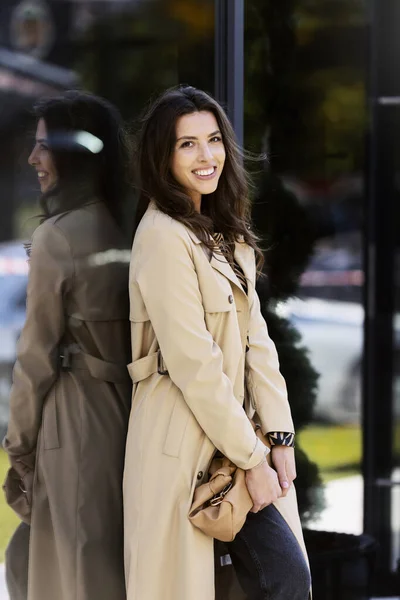 stock image Smiling, positive girl on the street, standing with her back to the glass building, leaning. A young woman in a beige, stylish coat.