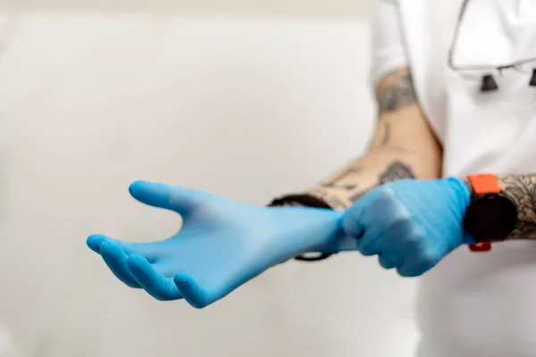 On a white background, a man with tattooed hands puts on disposable gloves. The concept of protection and safe work with the patient.