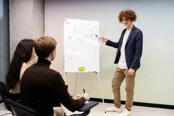 Smiling, positive guy points to graph drawn on flip chart. Young coach gives lecture to students