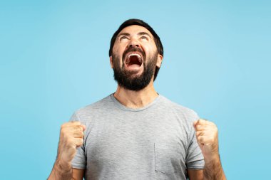 Sakallı adam zaferi kutluyor, yumruklarını kaldırıyor ve coşkulu mavi bir arka planda sevinçle çığlık atıyor, yoğun heyecan ve mutluluğu ifade ediyor.
