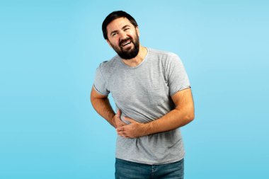 Sakallı bir adam mide ya da karın ağrısı çekiyor, iki eliyle karnını tutuyor, hazımsızlık çekiyor ya da diğer sindirim sorunları yaşıyor.