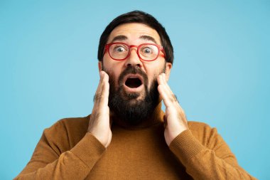 Sakallı ve kırmızı gözlüklü şaşırmış bir adam elleriyle yanaklarını tutuyor, mavi arka planda şaşkınlığını ve şaşkınlığını ifade ediyor.