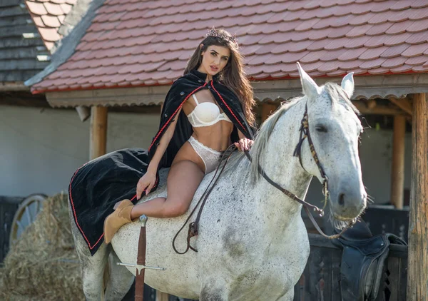 セクシーな白いビキニを着た官能的なブルネットの王女が馬に乗っています 岬と王冠と彼女の馬を持つ少女の肖像画 物語のおとぎ話のスタイルで馬と対話し 楽しみを持っている女の子 — ストック写真